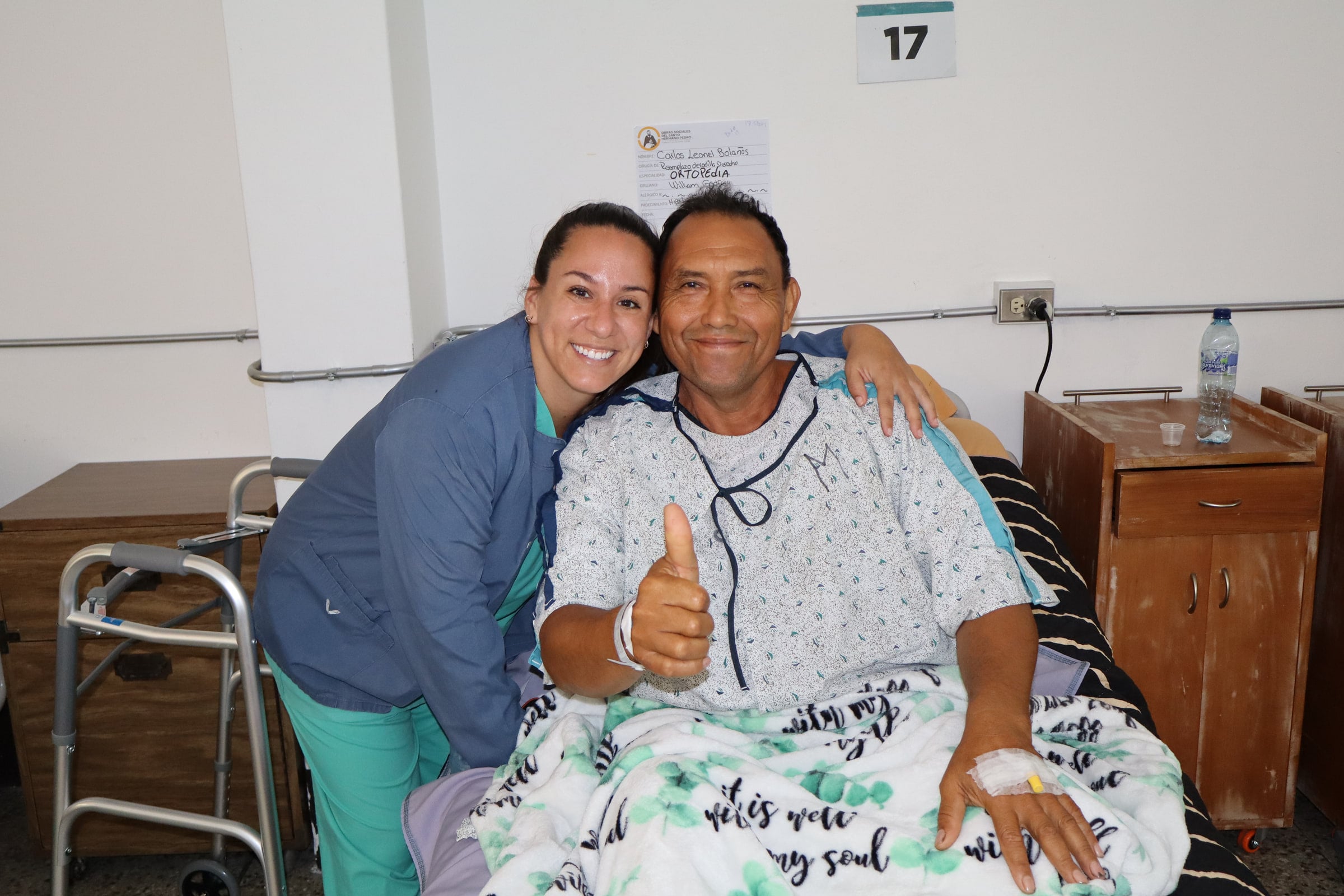 A patient in a hospital bed gives a thumbs up beside a smiling team member.