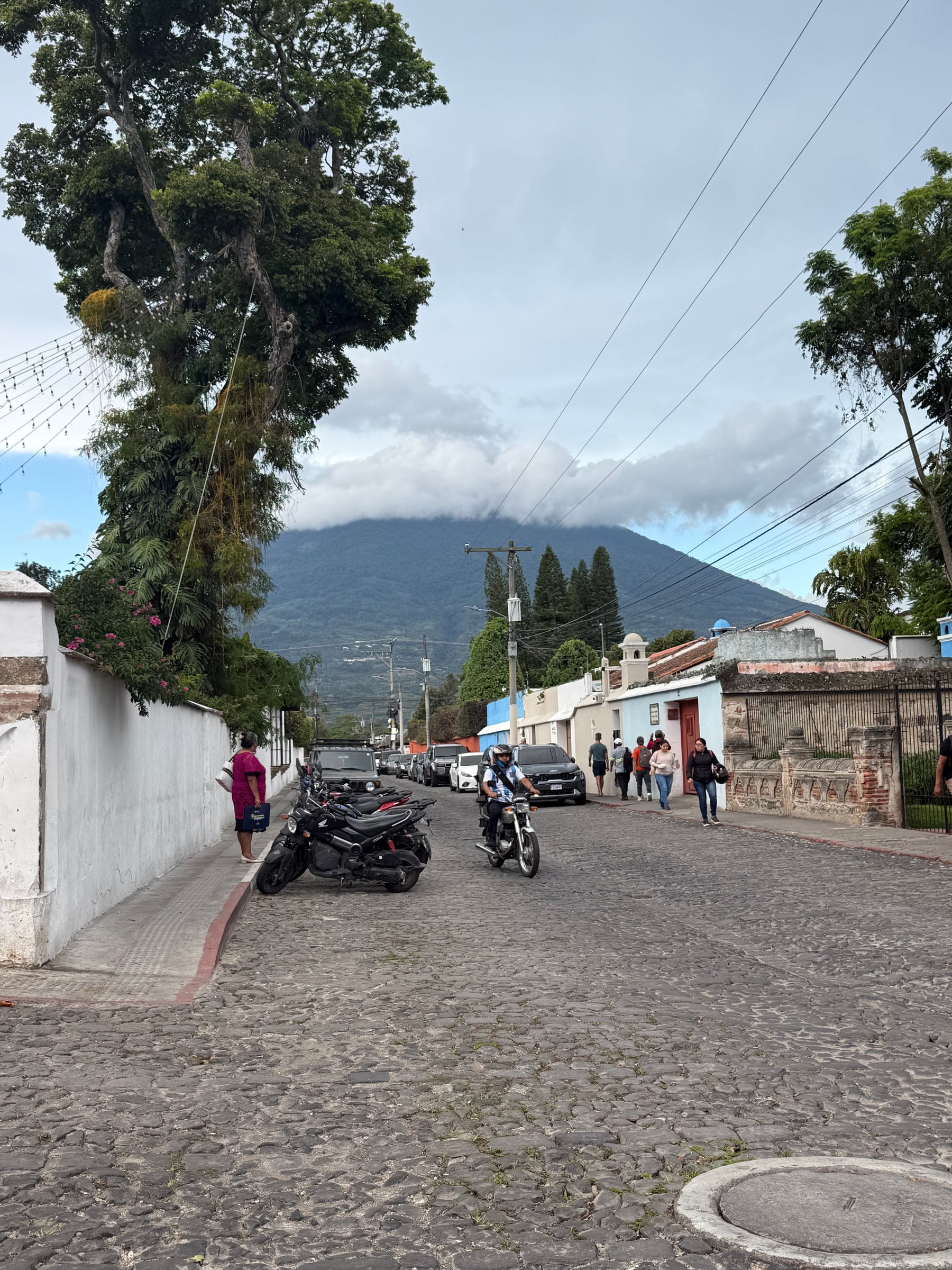 A cobblestone street in Antigua leads toward a volcano under a cloudy sky.