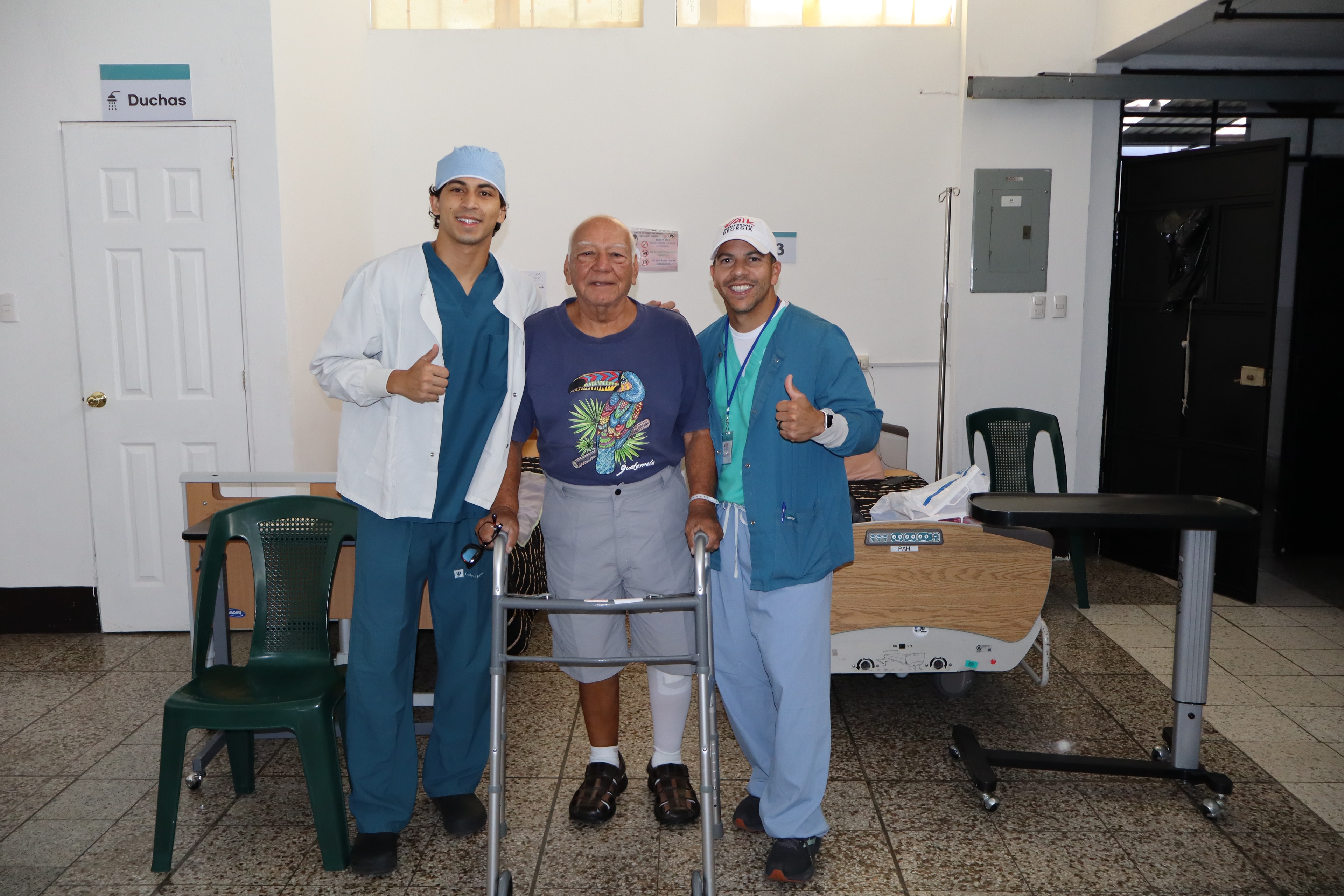 A smiling patient stands with a walker between two team members giving a thumbs up.