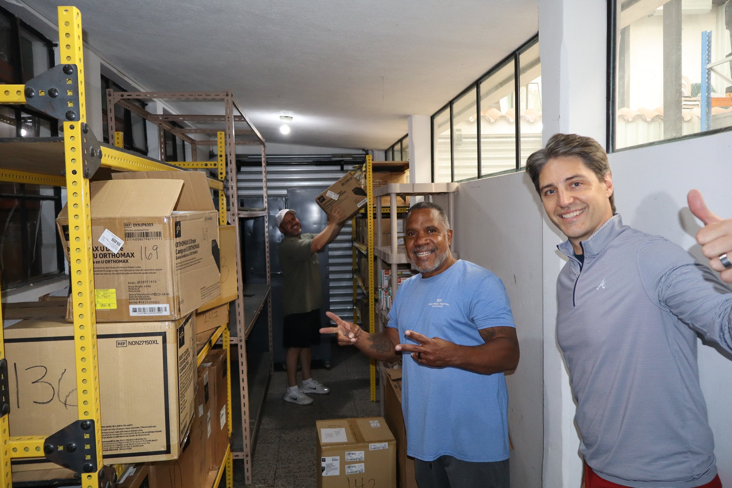 Two smiling team members stand among stacked supply boxes in a storeroom.
